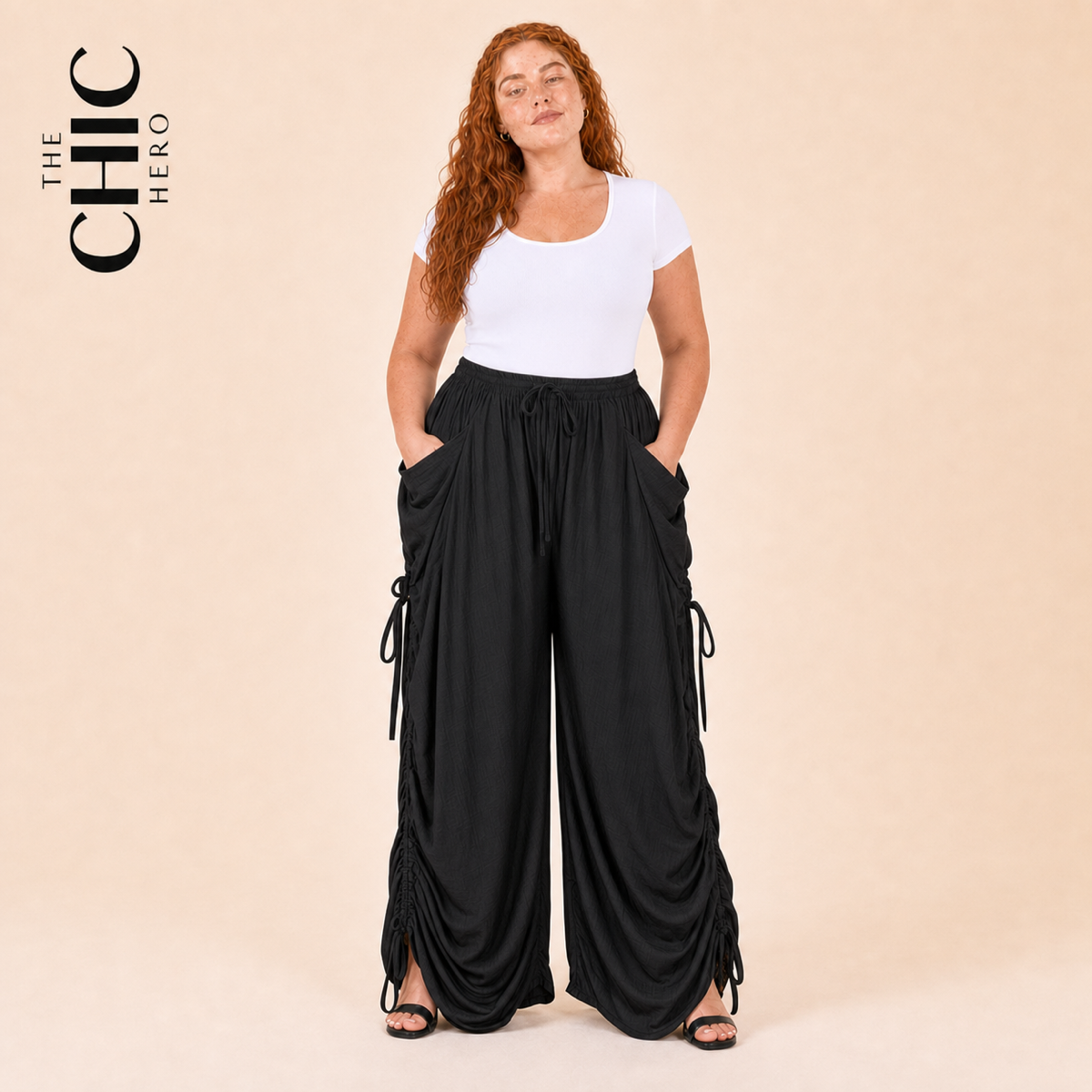 A woman modeling a white short-sleeve top and black draped wide-leg cargo pants with side tie details, posed against a solid beige studio background. The brand logo 'THE CHIC HERO' is visible in the top left corner.