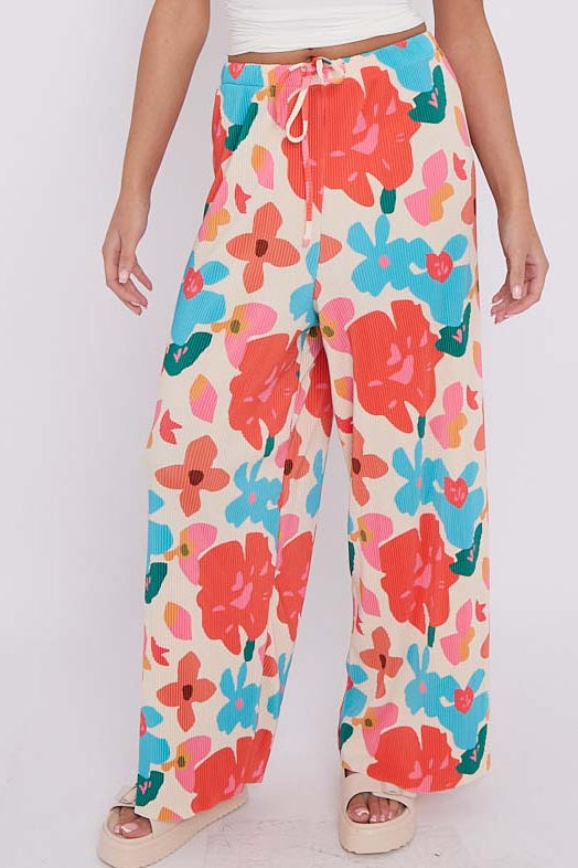 A model wearing wide-leg pants with a colorful floral print in orange, blue, and pink on an off-white background, paired with a coral orange top. The brand logo 'THE CHIC PARIS' is visible in the top left corner.