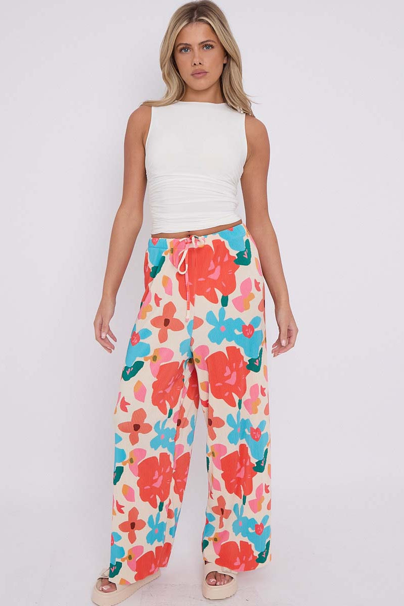 A model wearing wide-leg pants with a colorful floral print in orange, blue, and pink on an off-white background, paired with a coral orange top. The brand logo 'THE CHIC PARIS' is visible in the top left corner.