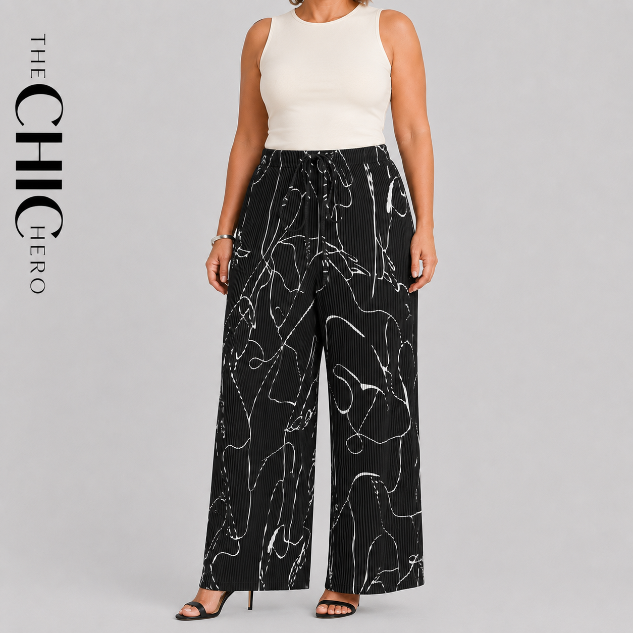 A model wearing black wide-leg pleated pants with a white abstract line pattern, paired with a cream sleeveless top, shown against a light gray studio background with 'THE CHIC HERO' text on the left.
