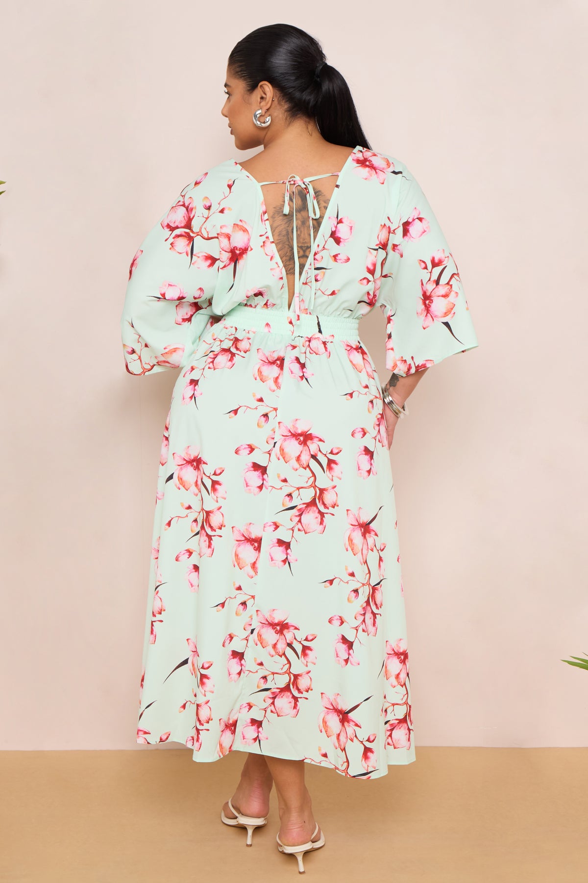 Woman wearing a floral dress standing against a beige wall with plants on either side.