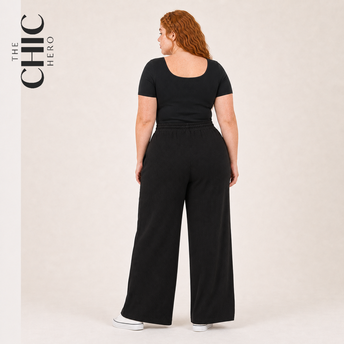 A woman with red hair is shown from the back wearing a black short-sleeve top and matching black wide-leg pants, standing against a plain light beige studio background. The brand name 'THE CHIC HERO' is printed vertically on the left side of the image.