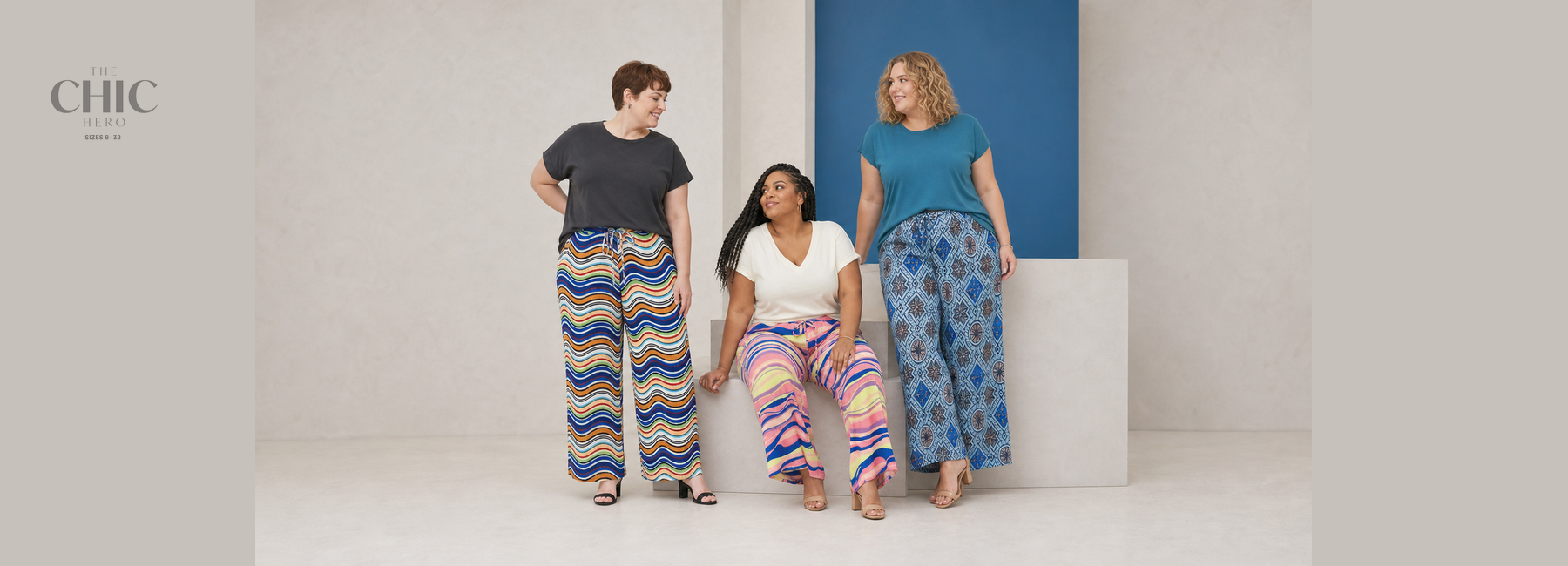 Three plus-size women wearing colourful pleated wide-leg trousers featuring wavy stripes, abstract prints and blue geometric patterns with simple tops.
