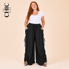 A woman modeling a white short-sleeve top and black draped wide-leg  cargo pants with side tie details, posed against a solid beige studio background. The brand logo 'THE CHIC HERO' is visible in the top left corner.