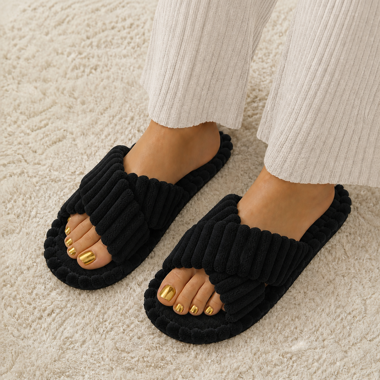 A person wearing black ribbed slide slippers with wide crisscross straps, paired with white ribbed pants, standing on a textured cream rug.