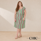 A woman modeling a sleeveless midi dress with vertical green, red, and white stripes, standing against a beige studio backdrop. The brand logo 'THE CHIC HERO' is visible in the bottom right corner.