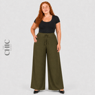 A woman modeling olive green wide-leg pinstripe pants with a drawstring waist, paired with a black short-sleeve top and black shoes, against a light gray studio background. The brand name 'The Chic Hero' is visible on the left side.