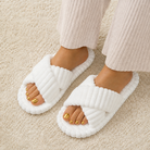 A person wearing white ribbed open-toe slippers with wide crisscross straps, standing on a beige textured rug. The person is also wearing light beige ribbed pants, and the toenails are painted gold.