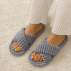 A person wearing gray ribbed open-toe slippers with crisscrossing straps, standing on a cream-colored textured rug. The person is also wearing cream ribbed pants.