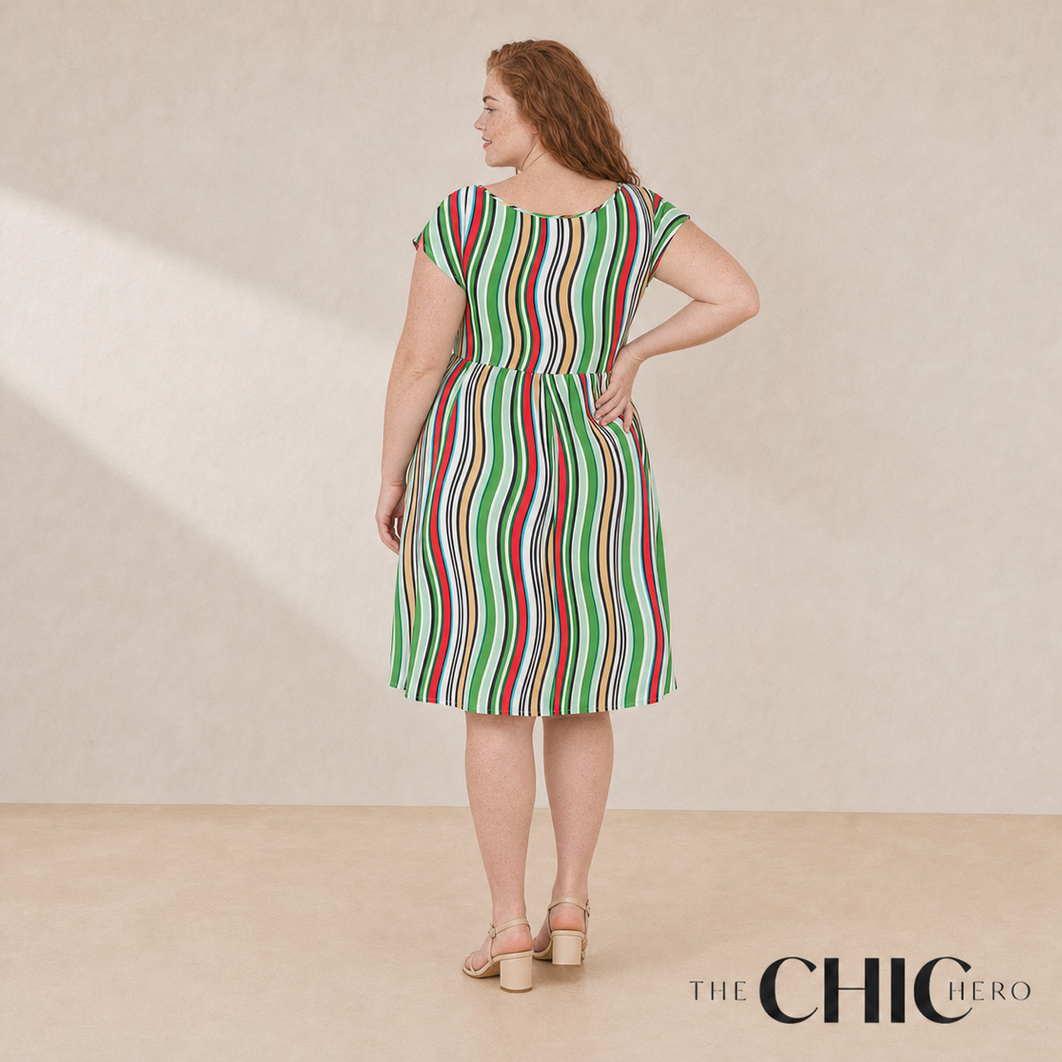 A woman stands with her back to the camera wearing a knee-length dress with vertical green, red, and white stripes. The photo is taken in a studio with a warm beige background, and the text 'THE CHIC HERO' appears in the bottom right corner.
