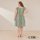 A woman stands with her back to the camera wearing a knee-length dress with vertical green, red, and white stripes. The photo is taken in a studio with a warm beige background, and the text 'THE CHIC HERO' appears in the bottom right corner.