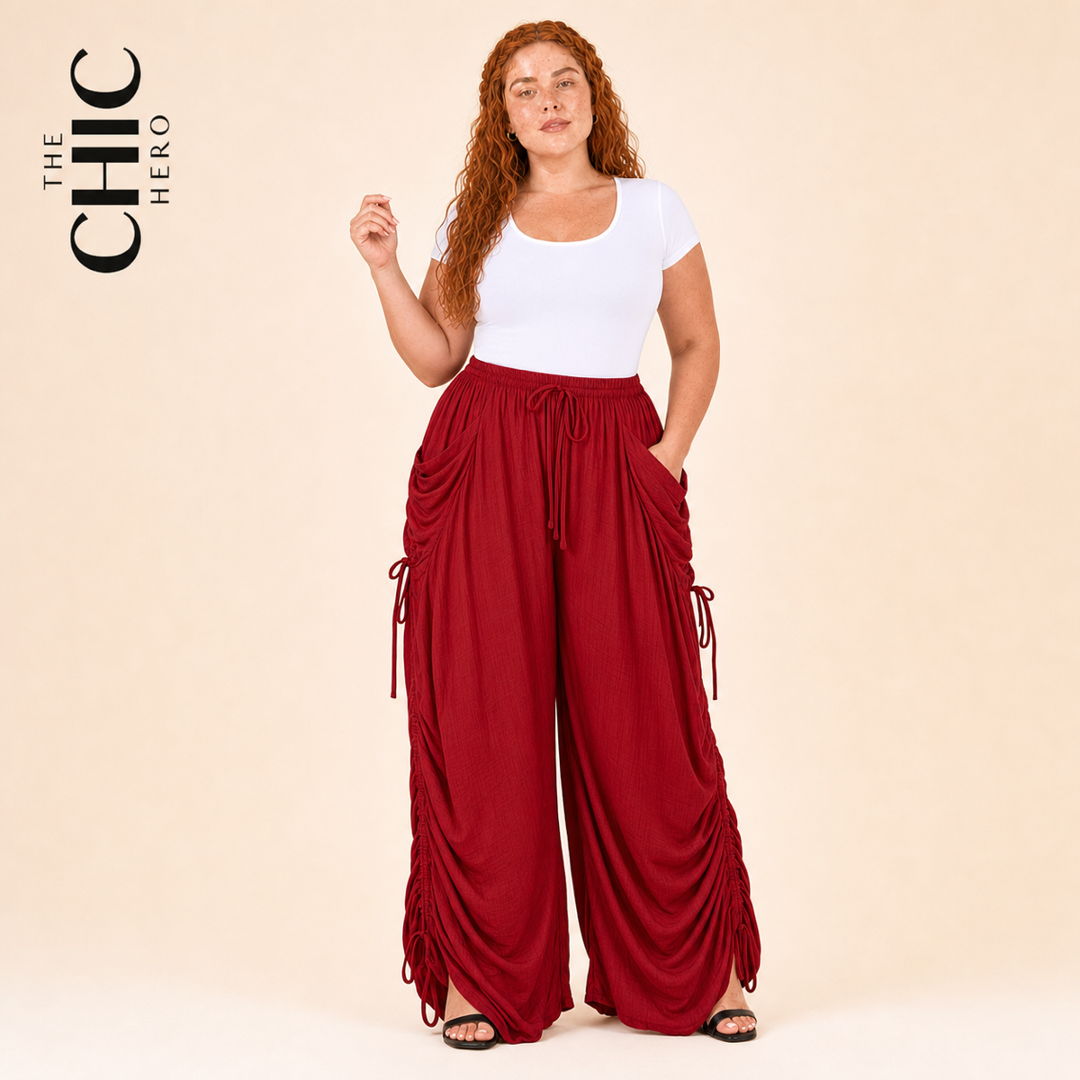 A woman modeling a white short-sleeve top and red draped wide-leg cargo pants with side tie details, posed against a solid beige studio background. The brand logo 'THE CHIC HERO' is visible in the top left corner.