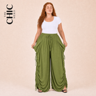 A woman modeling a white short-sleeve top and moss green draped wide-leg pants with side tie details, posed against a solid beige studio background. The brand logo 'THE CHIC HERO' is visible in the top left corner.