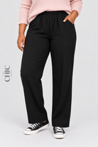 Women's plus size black stretch straight-leg trousers styled with a pale pink jumper and trainers