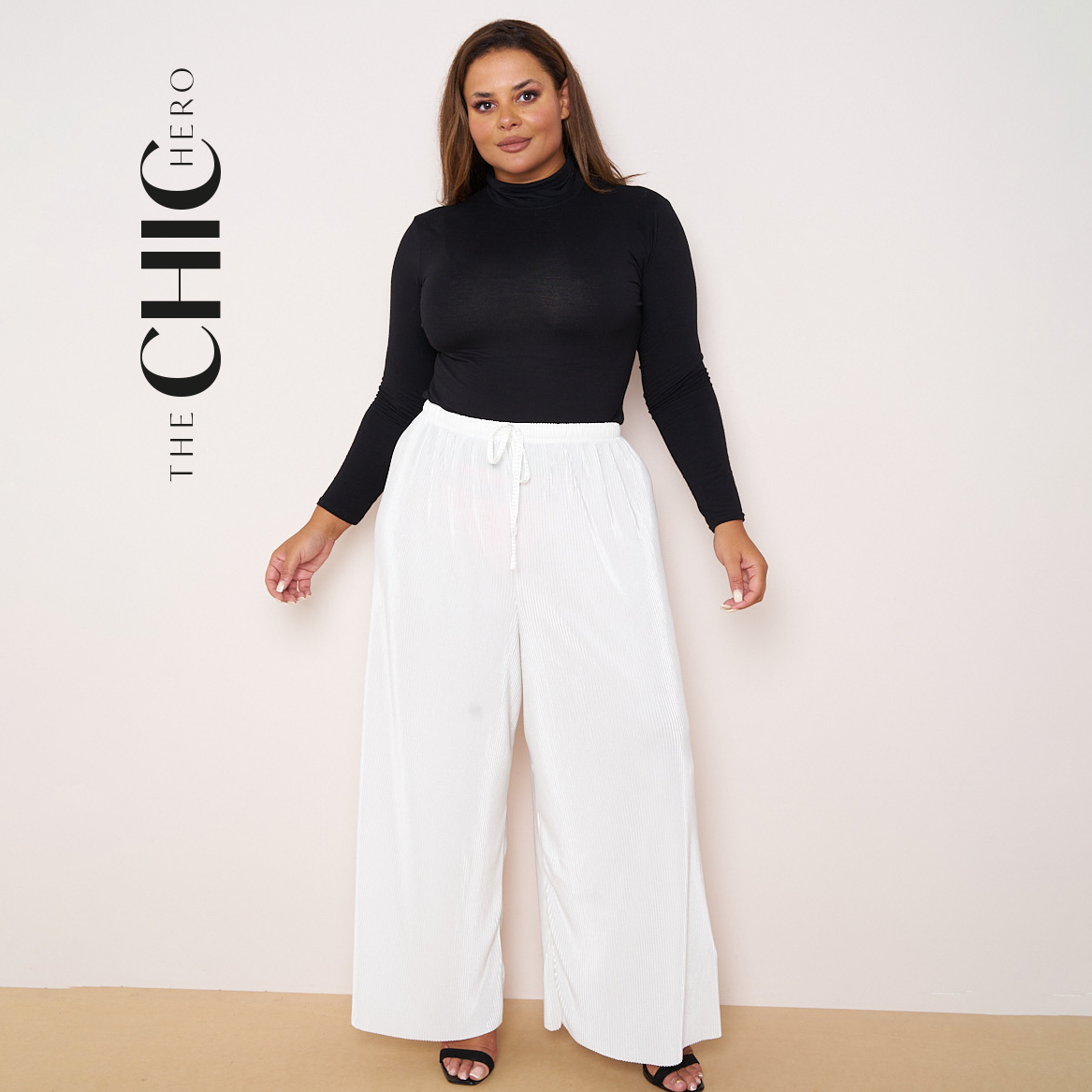 A model wearing white wide-leg drawstring pants and a black tank top, standing against a light gray studio background. The brand name 'THE CHIC HERO' is printed vertically on the left side.