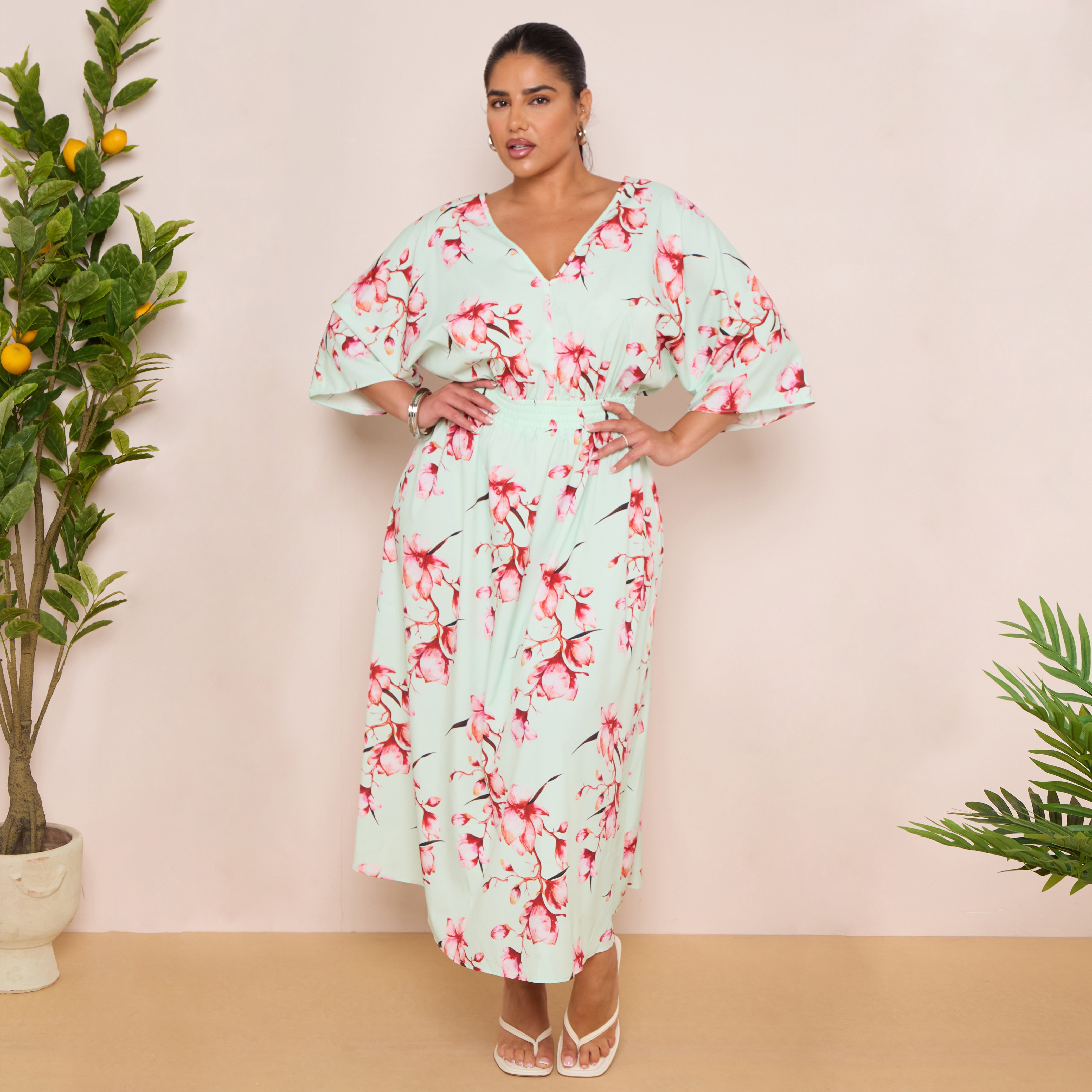 Woman wearing a floral dress standing in front of a pink wall with plants.