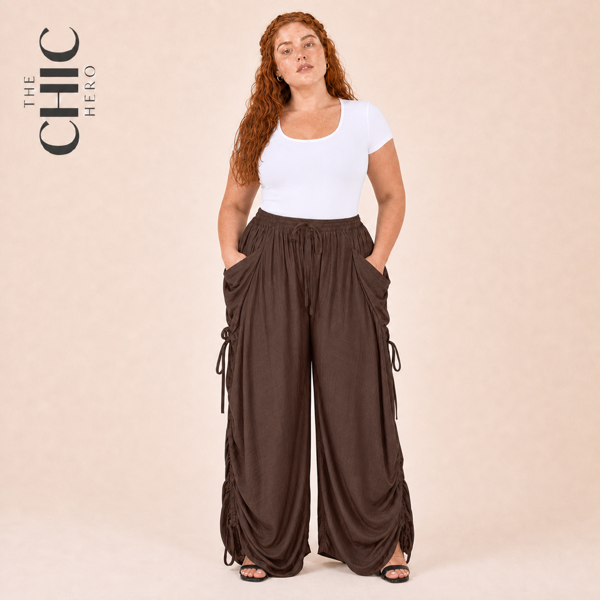 A woman modeling wide-leg brown cargo pants with side drawstrings, paired with a white t-shirt, standing against a solid beige background. The brand logo 'THE CHIC HERO' is visible in the top left corner.