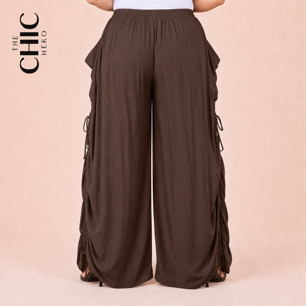 A person wearing wide-leg brown pants with side lace-up details, shown from the waist down against a light peach background. The brand logo 'THE CHIC HERO' is visible in the top left corner.