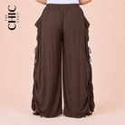 A person wearing wide-leg brown pants with side lace-up details, shown from the waist down against a light peach background. The brand logo 'THE CHIC HERO' is visible in the top left corner.