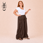 A woman modeling a white short-sleeve top and dark brown draped wide-leg pants with side tie details, posed against a solid beige studio background. The brand logo 'THE CHIC HERO' is visible in the top left corner.