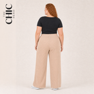 A woman shown from the back wearing beige wide-leg trousers and a black short-sleeve top, standing against a plain light beige studio background. The brand name 'THE CHIC HERO' appears vertically on the left side.