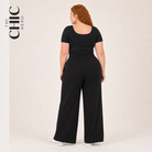 A woman with red hair is shown from the back wearing a black short-sleeve top and matching black wide-leg pants, standing against a plain light beige studio background. The brand name 'THE CHIC HERO' is printed vertically on the left side of the image.