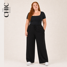 A woman modeling a black scoop-neck short-sleeve top and black wide-leg ribbed drawstring pants, standing against a plain beige studio background. The text 'THE CHIC HERO' appears in the upper left corner.