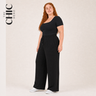 A woman modeling a black short-sleeve jumpsuit with an asymmetrical neckline, drawstring waist, and wide legs, standing against a light beige studio background. The brand name 'THE CHIC HERO' is printed vertically on the left side of the image.