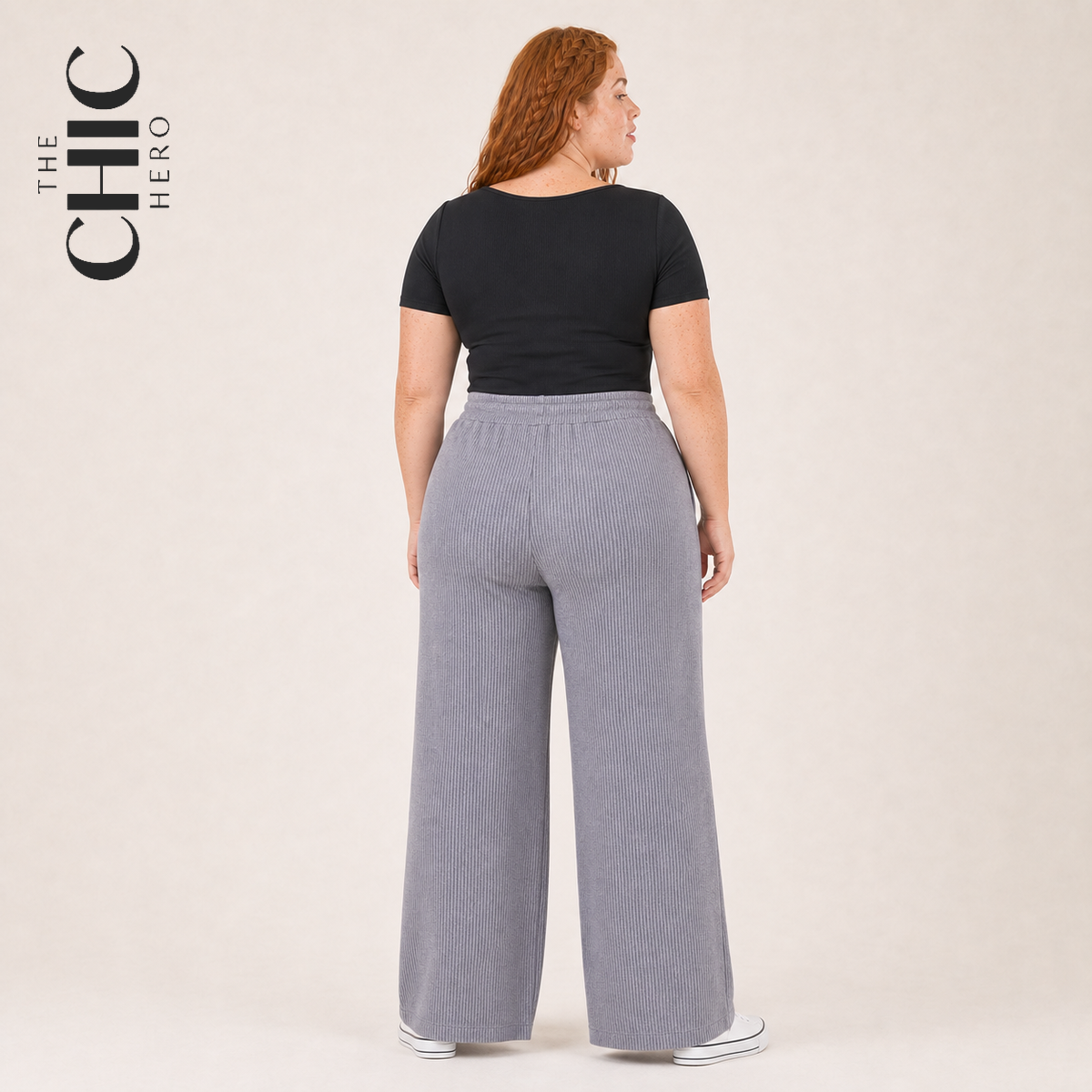 A woman shown from the back wearing a black short-sleeve top and light gray wide-leg ribbed pants, standing against a plain light beige studio background. The brand name 'THE CHIC HERO' appears in the upper left corner.