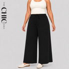 A model wearing black wide-leg drawstring pants with a white ribbed tank top and white loafers, shown against a light gray studio background. The brand name 'THE CHIC HERO' is printed vertically on the left side.