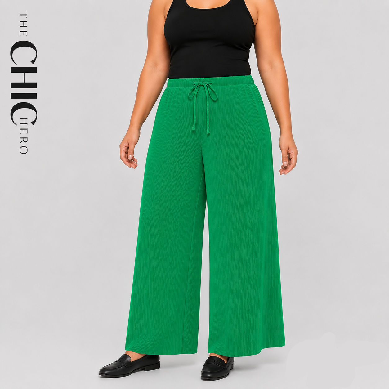 A model wearing bright green wide-leg drawstring pants with a black tank top and black loafers, shown against a light gray studio background with 'THE CHIC HERO' branding.