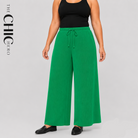 A model wearing bright green wide-leg drawstring pants with a black tank top and black loafers, shown against a light gray studio background with 'THE CHIC HERO' branding.