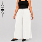 A model wearing white wide-leg drawstring pants and a black tank top, standing against a light gray studio background. The brand name 'THE CHIC HERO' is printed vertically on the left side.