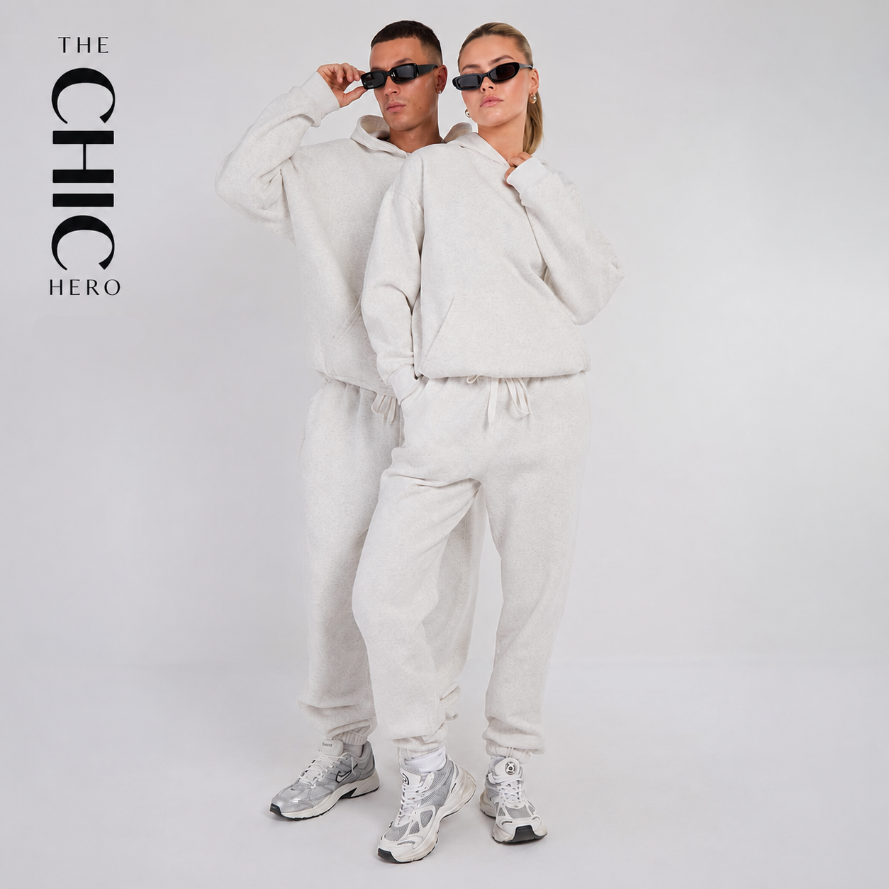 A man and a woman modeling matching light beige/cream hoodies and jogger sweatpants sets against a plain light gray studio background. The woman is in front wearing sunglasses, and the man is behind wearing sunglasses.