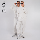 A man and a woman modeling matching light beige/cream hoodies and jogger sweatpants sets against a plain light gray studio background. The woman is in front wearing sunglasses, and the man is behind wearing sunglasses.